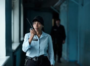 Young female security guard in uniform and cap speaking on walkie-talkie to her colleague while moving along corridor of building